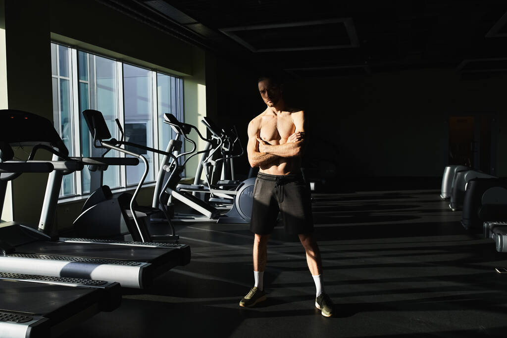 Muskularny mężczyzna bez koszuli stojący pewnie przed rzędem bieżnie w siłowni. - Zdjęcie, obraz