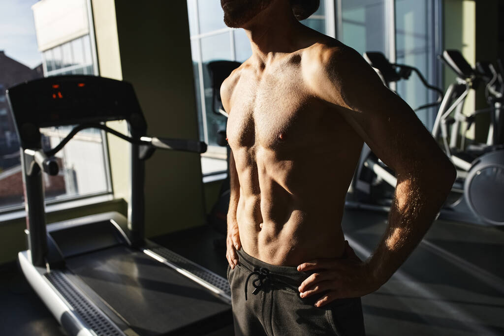 Ein muskulöser Mann ohne Hemd steht selbstbewusst neben einem Laufband in einem Fitnessstudio. - Foto, Bild
