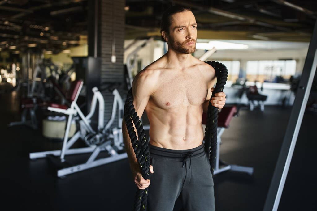 Без рубашки, мускулистый человек интенсивно сосредоточен на тренировке в спортзале с тяжелыми канатами. - Фото, изображение