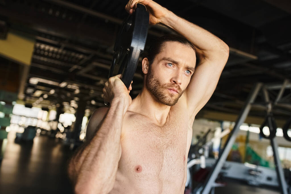 Egy félmeztelen férfi megragad egy súlyzót egy edzőteremben, bemutatva izmos testalkatát, miközben az edzésprogramjára koncentrál.. - Fotó, kép