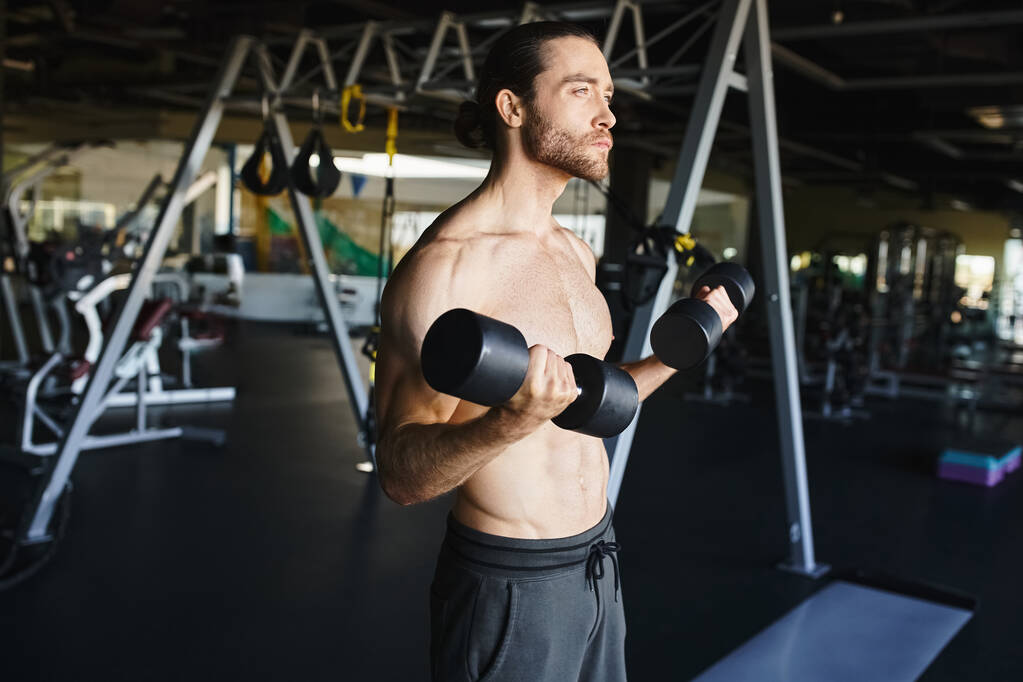 Un hombre musculoso en un gimnasio, sin camisa, sostiene con confianza dos pesas, mostrando su dedicación al entrenamiento de fuerza. - Foto, imagen