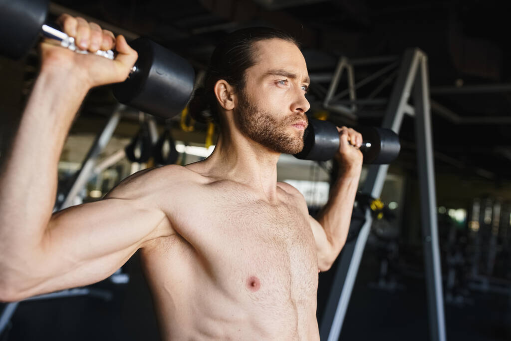 Un hombre sin camisa empujándose hasta sus límites, sosteniendo pesas en un gimnasio. - Foto, imagen