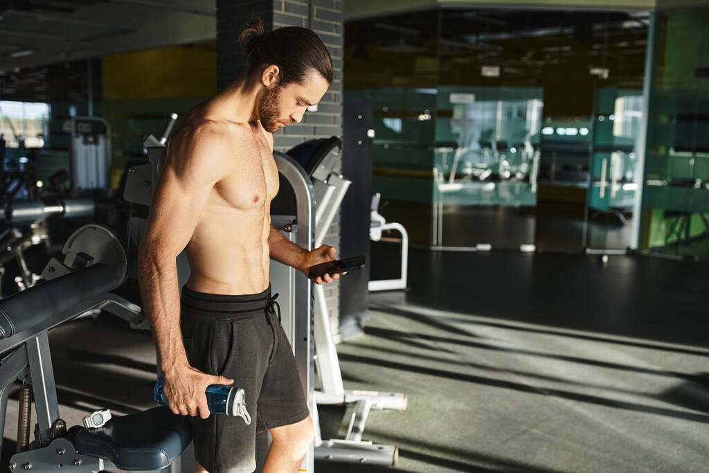 Hombre sin camisa en el gimnasio, intensamente enfocado en la pantalla del teléfono celular mientras hace ejercicio. - Foto, Imagen