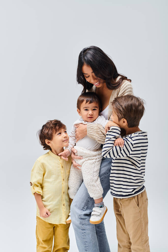 Asiatische Mutter hält ihr Baby zärtlich in der Hand, während zwei kleine Kinder zusehen und einen herzerwärmenden Familienmoment in einem Atelier schaffen. - Foto, Bild