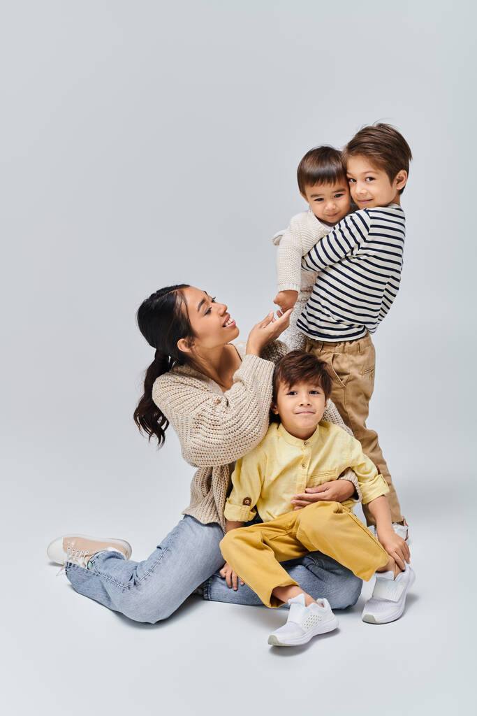 Una joven madre asiática se sienta en el suelo, abrazando calurosamente a sus hijos en un ambiente de estudio con un fondo gris. - Foto, imagen