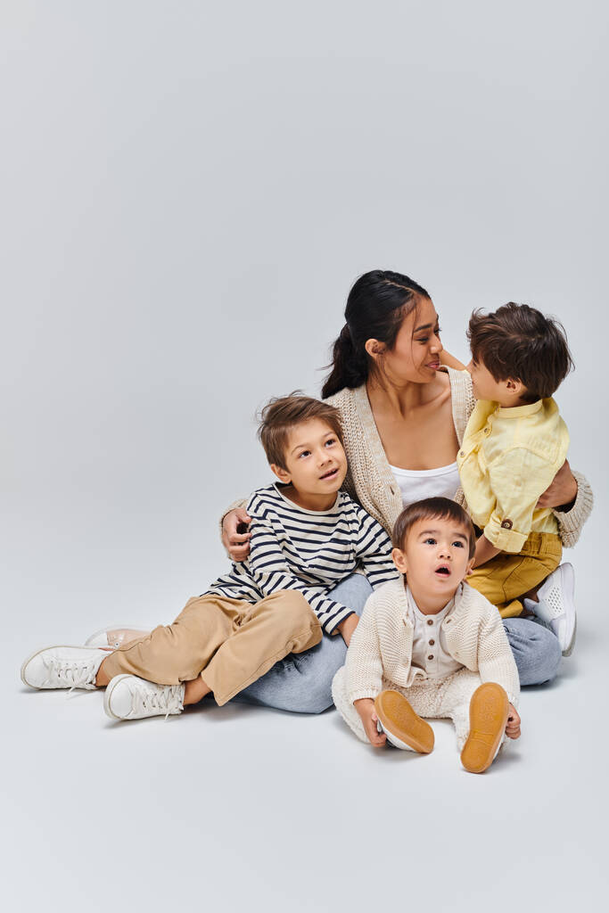 A young Asian mother sitting on the ground with her children in a serene studio setting on a grey background. - Photo, Image