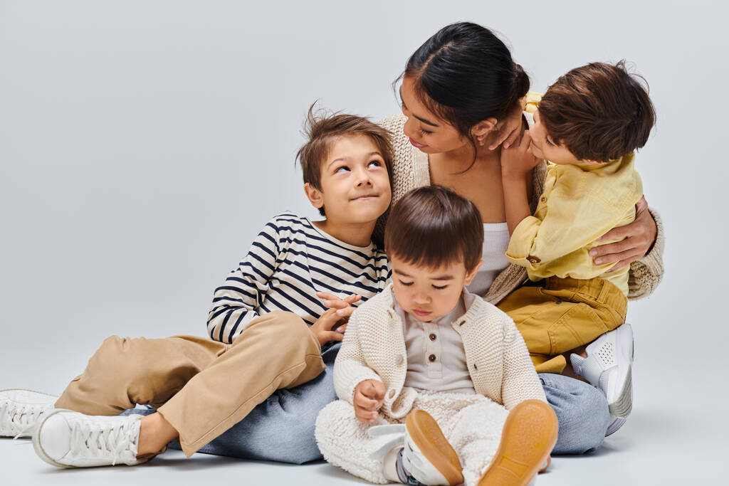 Eine asiatische Mutter sitzt mit ihren Kindern auf dem Boden und umarmt sich in einem Studio vor grauem Hintergrund liebevoll. - Foto, Bild