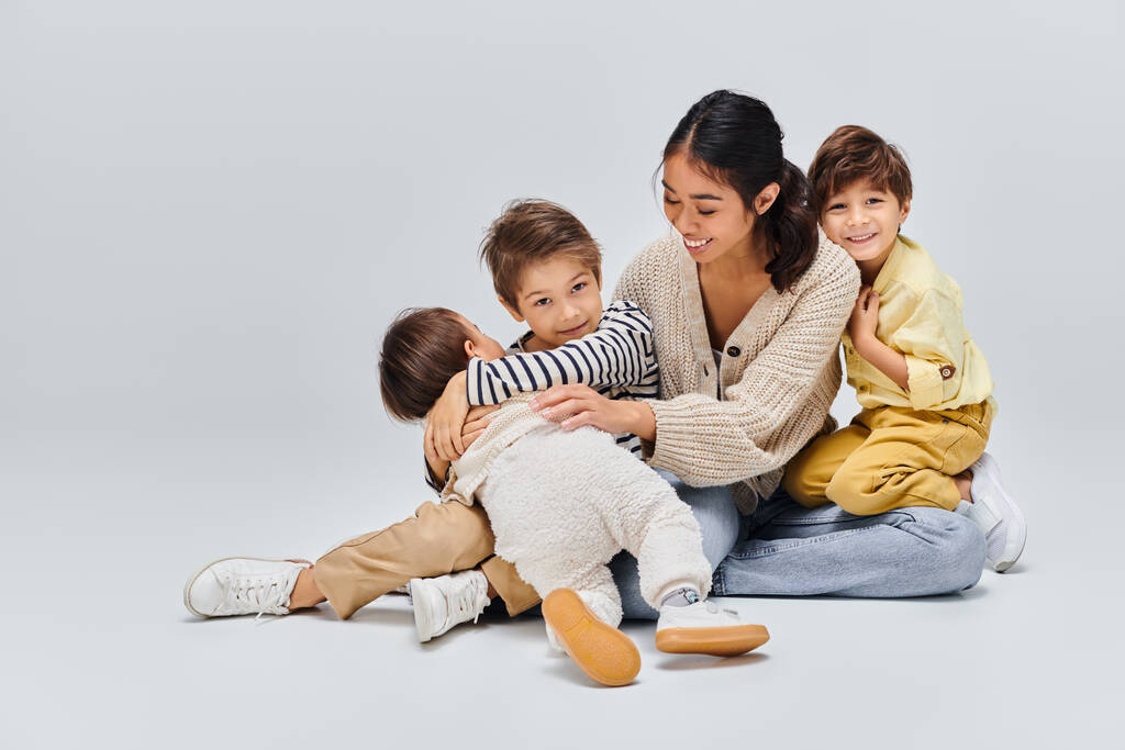 Eine junge asiatische Mutter sitzt auf dem Boden, umgeben von ihren Kindern, und teilt einen Moment der Liebe und Nähe. - Foto, Bild