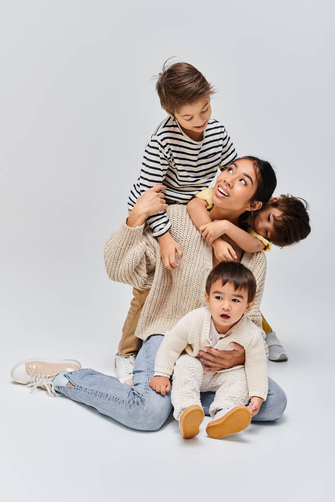 Una giovane madre asiatica e i suoi figli si siedono uno sopra l'altro, creando una piramide umana in uno studio su uno sfondo grigio. - Foto, immagini