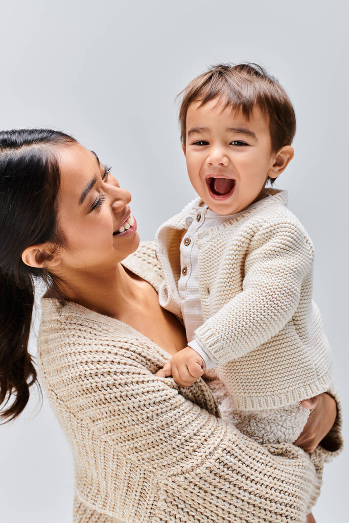 Une jeune mère asiatique tient tendrement son bébé dans ses bras dans un décor de studio sur fond gris. - Photo, image