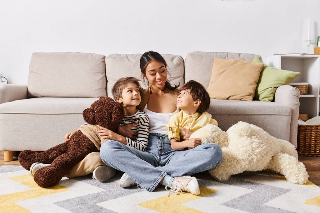 Mladá asijská matka sedí na podlaze se svými dvěma dětmi a medvídkem v obývacím pokoji. - Fotografie, Obrázek