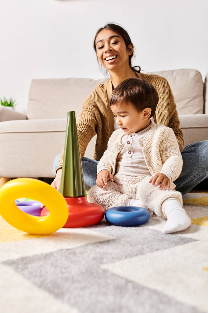 Eine junge asiatische Mutter sitzt auf dem Boden und spielt fröhlich mit ihrem kleinen Sohn im Wohnzimmer ihres Hauses. - Foto, Bild