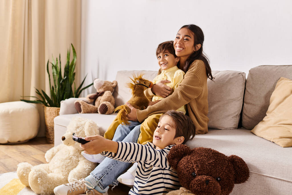 Μια νεαρή Ασιάτισσα μητέρα και οι γιοι της κάθονται σε έναν καναπέ περικυκλωμένοι από λούτρινα ζωάκια, δημιουργώντας μια ζεστή και οικεία σκηνή.. - Φωτογραφία, εικόνα
