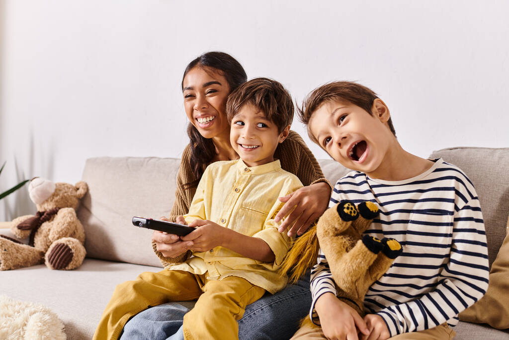 lapset, yhdessä aasialaisen äitinsä kanssa, rentoutua sohvalla pitämällä kaukosäätimiä viihtyisässä olohuoneessa. - Valokuva, kuva