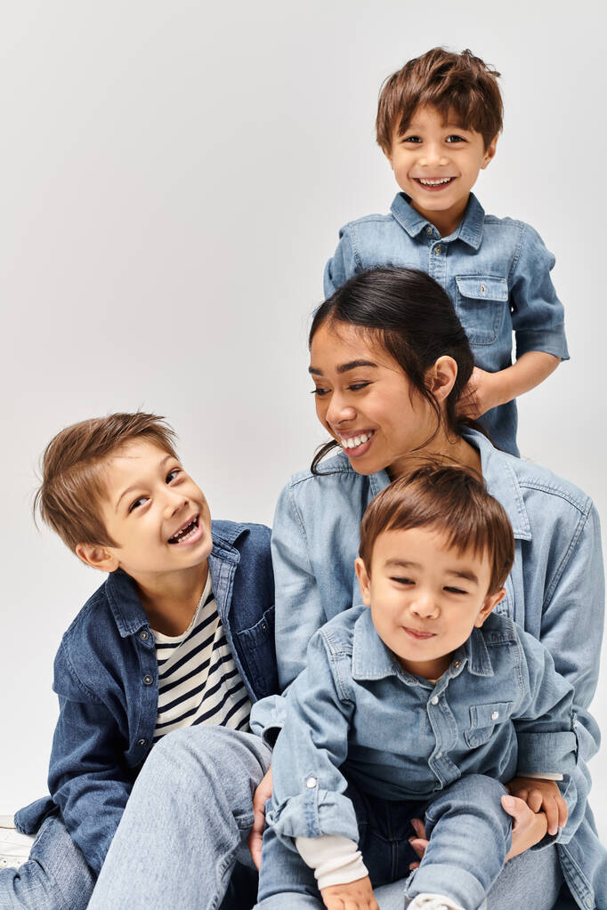 Μια ομάδα μικρών παιδιών, μια μητέρα, και οι γιοι της, ντυμένοι με τζιν ρούχα, κάθονται το ένα δίπλα στο άλλο σε ένα γκρι στούντιο.. - Φωτογραφία, εικόνα