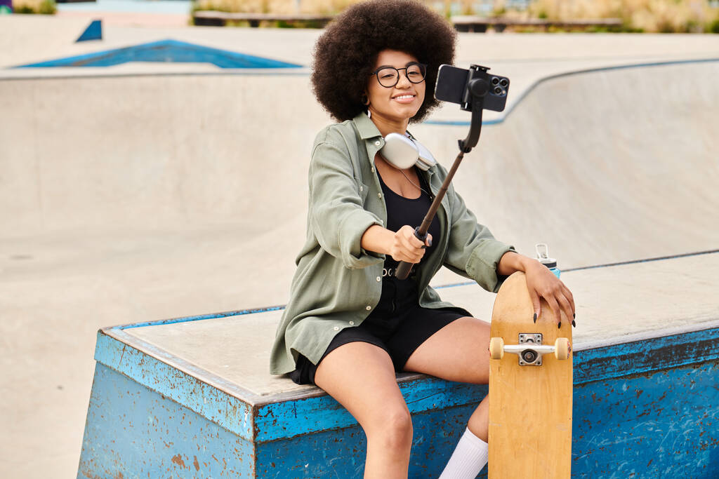 Молодая афроамериканка с вьющимися волосами сидит на синей скамейке, держа скейтборд на открытом скейт-парке.. - Фото, изображение