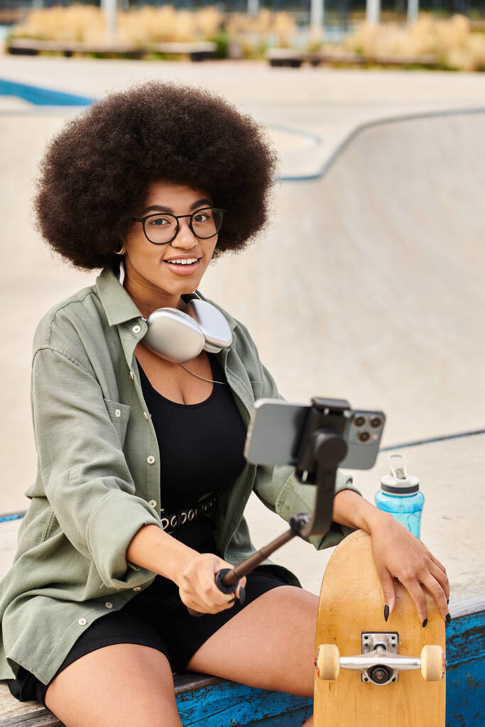 Молодая афроамериканка с вьющимися волосами сидит на выступе, держа селфи-палку в ярком скейт-парке. - Фото, изображение