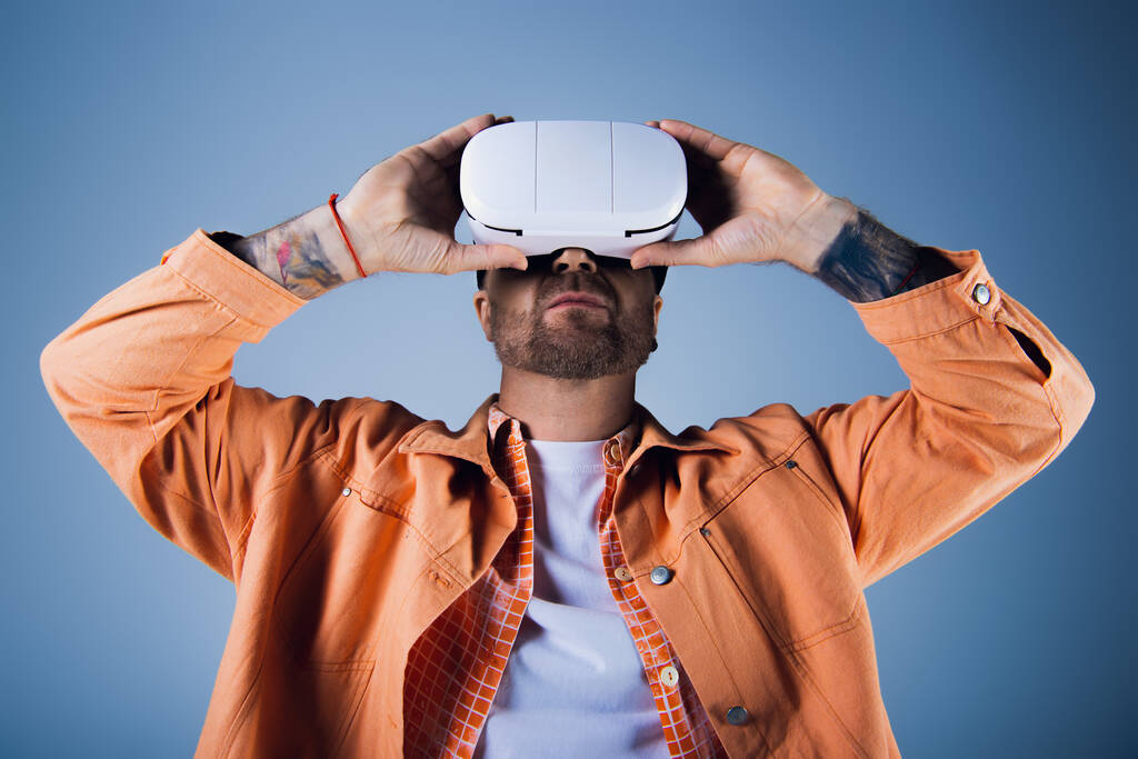 A man in an orange shirt raises a white vr headset above his head in a studio setting. - Photo, Image