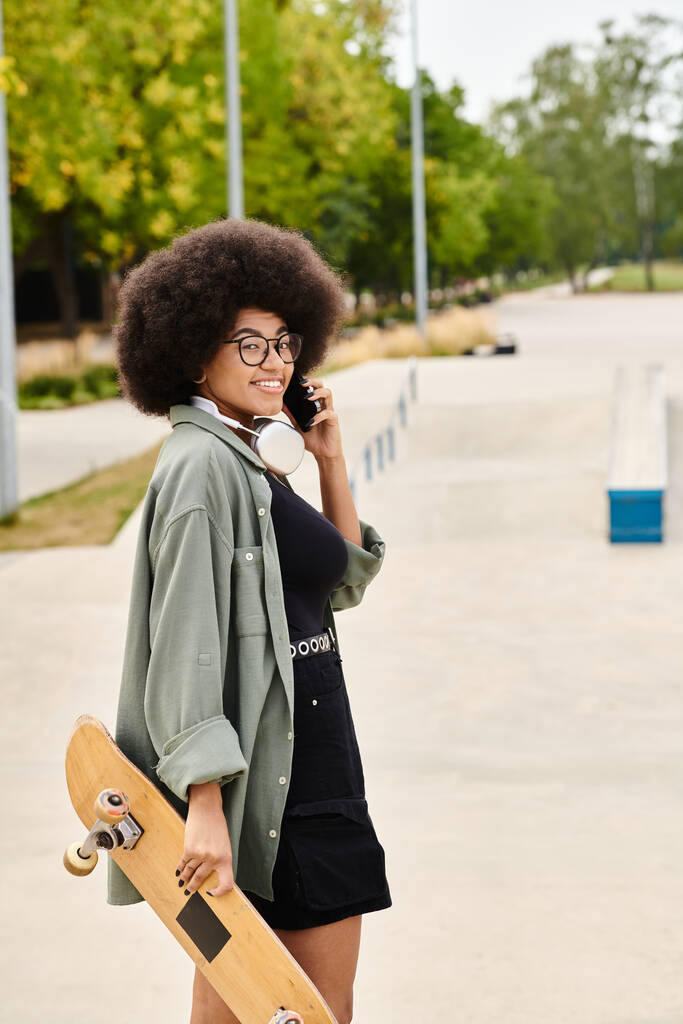 Egy fiatal afro-amerikai nő, göndör hajjal, többfeladatú, gördeszkát tart, és mobilon beszél egy gördeszkás parkban.. - Fotó, kép