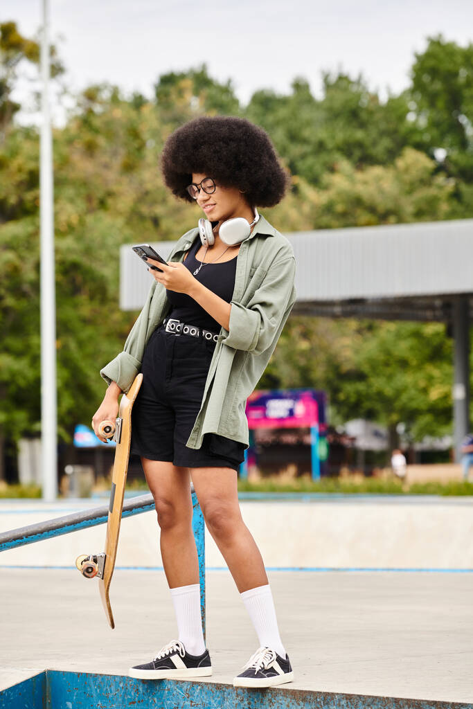 Uma jovem afro-americana com cabelo encaracolado confiantemente fica em uma borda com seu skate em um cenário de parque de skate. - Foto, Imagem