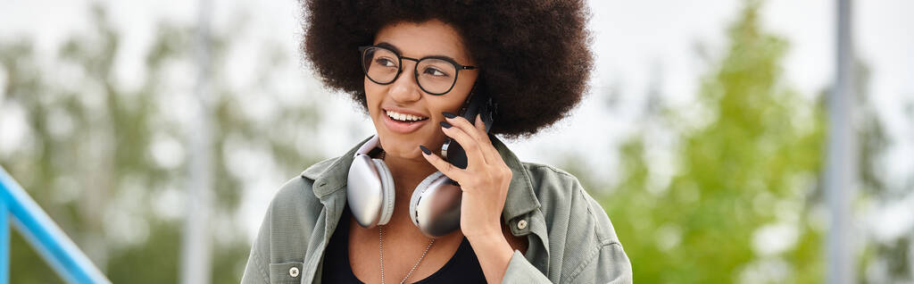 Eine stylische Frau mit lockigem Haar und Brille spricht auf ihrem Handy im Freien. - Foto, Bild