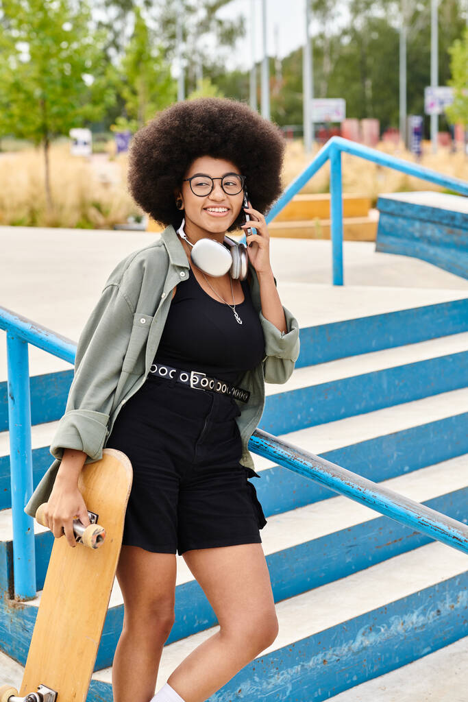 Una joven afroamericana con el pelo rizado sosteniendo un monopatín y hablando por celular en un parque de skate. - Foto, imagen