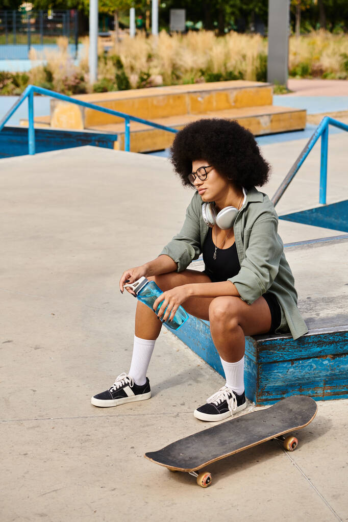 Młoda Afroamerykanka z kręconymi włosami siedząca elegancko na ławce trzymając butelkę wody w skateparku. - Zdjęcie, obraz