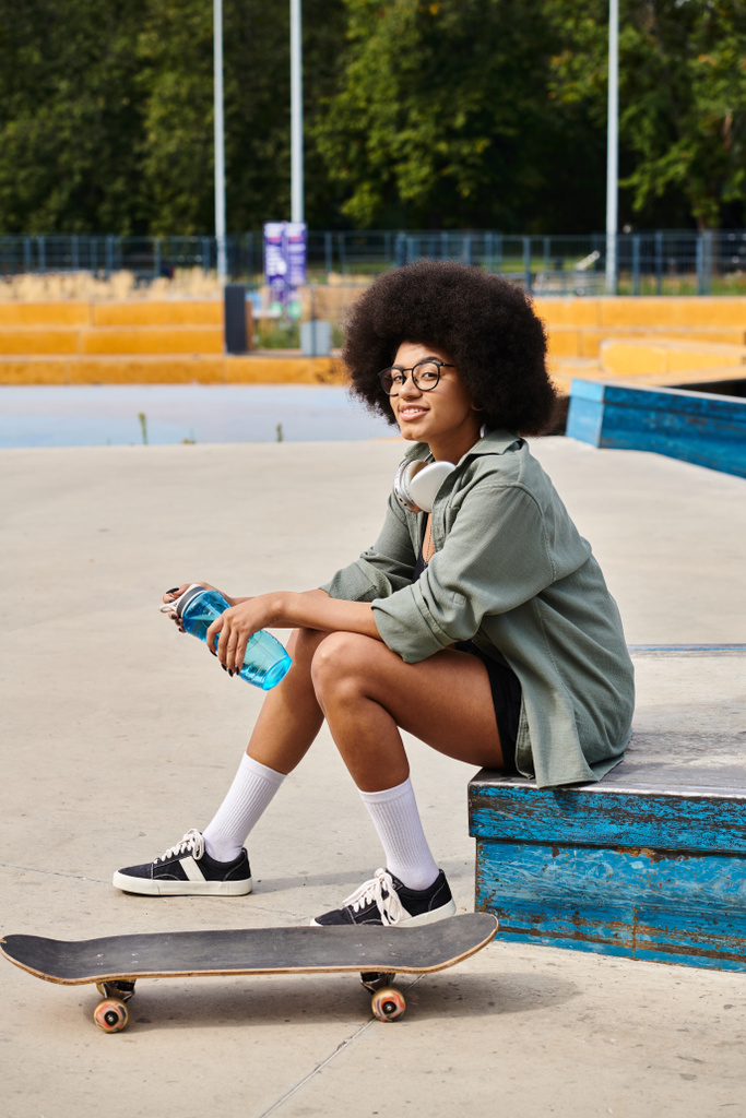 Jonge Afro-Amerikaanse vrouw met krullend haar zittend op een skateboard, met een fles water in een outdoor skate park. - Foto, afbeelding