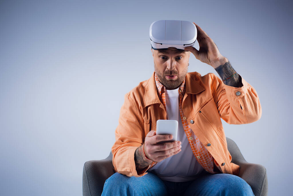 Un hombre en una silla sostiene un teléfono inteligente en un entorno de estudio, inmerso en el mundo de la metáfora a través de sus auriculares VR. - Foto, imagen