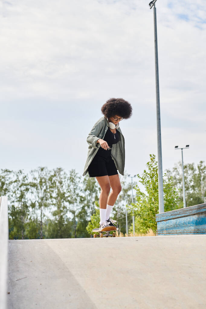 Μια νεαρή Αφρο-Αμερικανίδα με σγουρά μαλλιά καβαλάει με αυτοπεποίθηση ένα σκέιτμπορντ σε μια τσιμεντένια ράμπα σε ένα υπαίθριο πάρκο για σκέιτ.. - Φωτογραφία, εικόνα