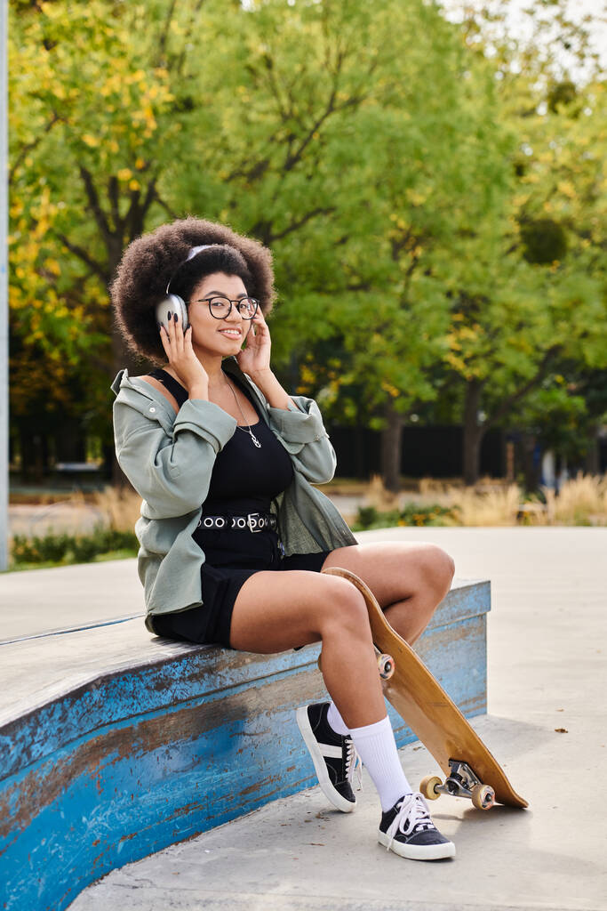 Eine junge Afroamerikanerin mit lockigem Haar sitzt auf einer Bank, vertieft in ein Gespräch auf ihrem Handy in einem Skatepark. - Foto, Bild