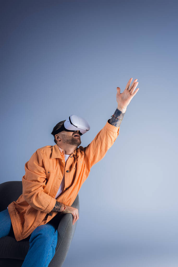 Een man in een VR-headset zit in een stoel met zijn hand omhoog, ondergedompeld in een virtuele wereld in een studio-omgeving. - Foto, afbeelding