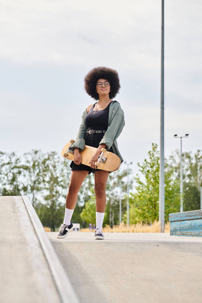 カーリーヘアの若いアフリカ系アメリカ人女性は自信を持ってスケートパークのランプの上のスケートボードに立っています. - 写真・画像