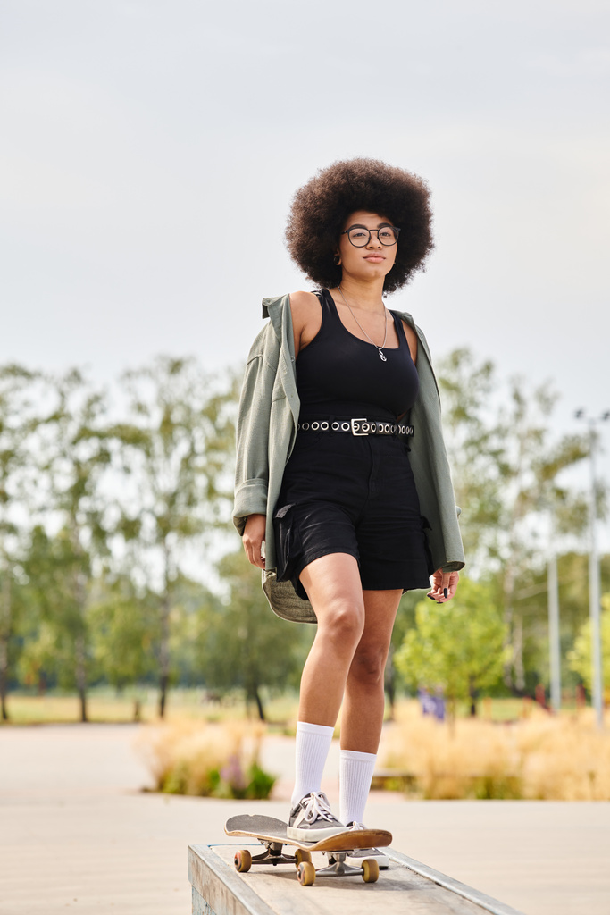 Μια νεαρή Αφρο-Αμερικανίδα στέκεται με αυτοπεποίθηση πάνω σε ένα σκέιτμπορντ σε ένα πάρκο, δείχνοντας τις ικανότητες και το στυλ της.. - Φωτογραφία, εικόνα