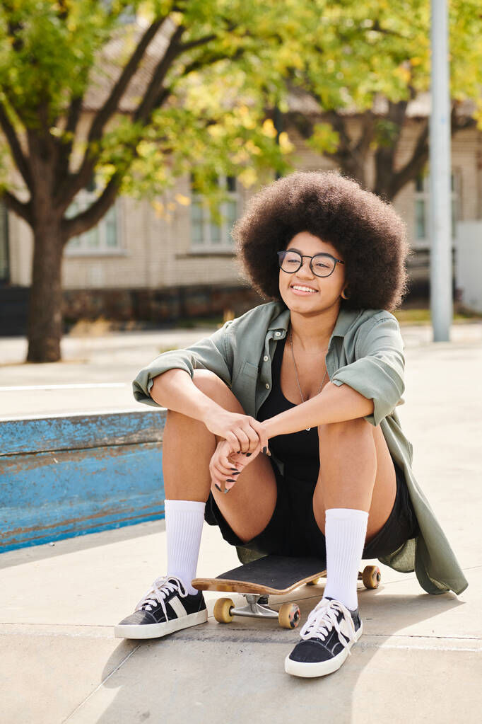 Une jeune femme afro-américaine élégante avec une coiffure afro volumineuse s'assoit tranquillement sur une planche à roulettes dans un parc de skate dynamique. - Photo, image