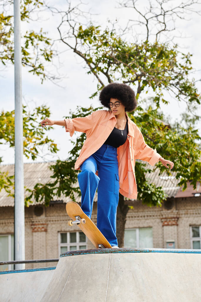 Una joven afroamericana con el pelo rizado monta hábilmente un monopatín al lado de una rampa en un parque de skate al aire libre. - Foto, imagen