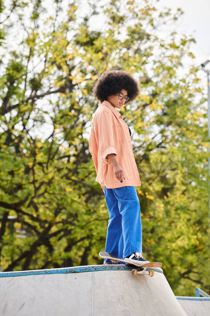 A curly-haired young man confidently rides a skateboard on top of a cement ramp in a skate park. - Photo, Image