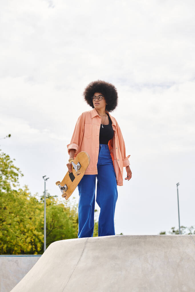 Une jeune Afro-Américaine avec une coiffure afro bouclée tient en toute confiance une planche à roulettes dans un skate park en plein air. - Photo, image
