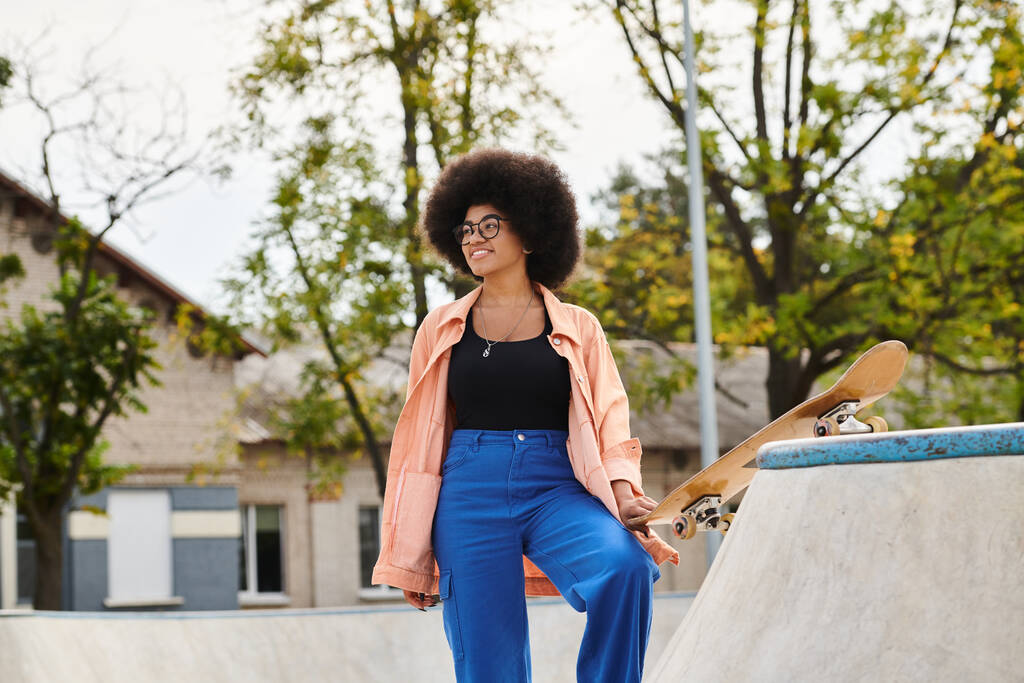 Young African American woman with curly hair stands next to skateboard on a skateboard ramp in a vibrant outdoor skate park. - Photo, Image