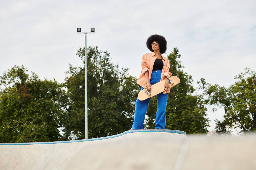 A young African American woman with curly hair confidently stands atop a skateboard ramp at an outdoor skate park. - Photo, Image
