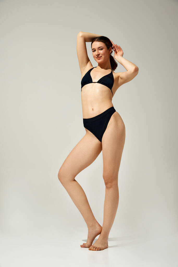 Pełnowymiarowy obraz pięknej młodej dziewczyny z szczupłym ciałem stojącej w czarnych strojach kąpielowych, pozującej na szarym tle studia. Pojęcie pielęgnacji ciała i zdrowia, kobieca uroda, wellness - Zdjęcie, obraz
