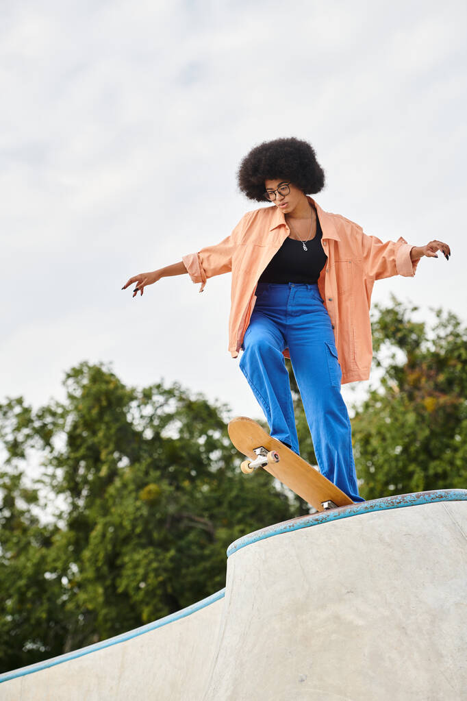 Egy göndör hajú afroamerikai nő magabiztosan gördeszkázik a rámpa oldalán egy szabadtéri gördeszka parkban.. - Fotó, kép