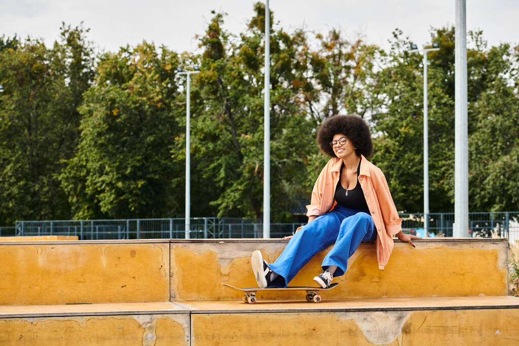 Egy fiatal afro-amerikai nő göndör hajjal magabiztosan ül a párkányon gördeszkával egy vibráló gördeszka parkban.. - Fotó, kép