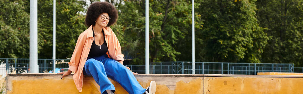 Nuori afroamerikkalainen nainen, jolla on kiharat hiukset, istuu kauniisti seinällä, ylittää jalkansa katsellen kaukaisuuteen.. - Valokuva, kuva