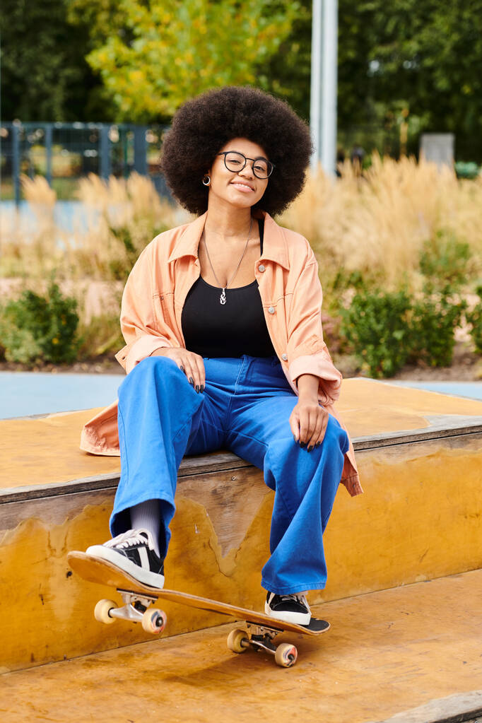 Eine junge Afroamerikanerin mit lockigem Haar sitzt auf einem Sims und hält ein Skateboard im Skatepark. - Foto, Bild
