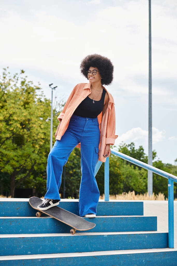 Μια Αφροαμερικανή γυναίκα με σγουρά μαλλιά skateboarding επιδέξια κάτω από μια σκάλα σε ένα αστικό πάρκο skate. - Φωτογραφία, εικόνα