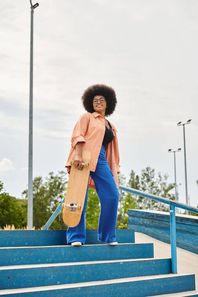 Een jonge Afro-Amerikaanse vrouw met krullend haar staat vol vertrouwen op de trap, met een skateboard in een stedelijk skatepark. - Foto, afbeelding