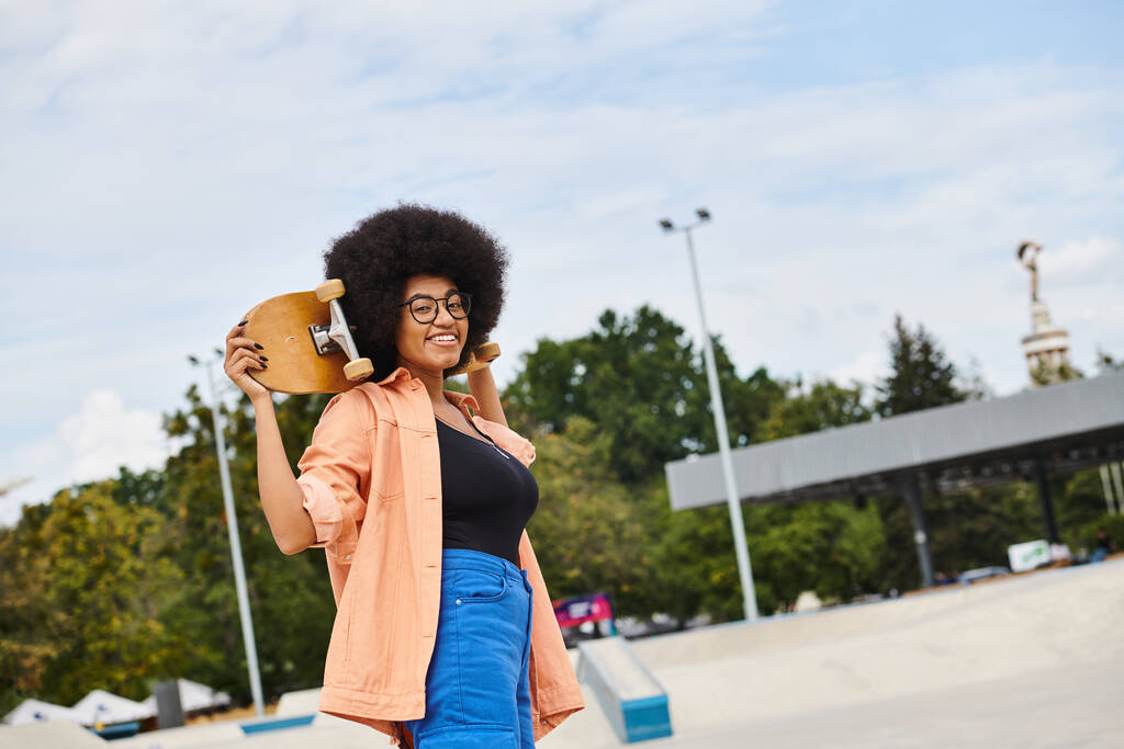 カーリーヘアのスタイリッシュなアフリカ系アメリカ人女性がスケートパークで右手にスケートボードを保持. - 写真・画像