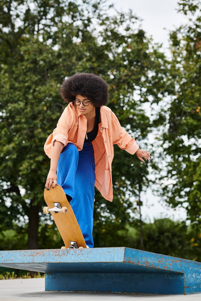 Mladá Afroameričanka s kudrnatými vlasy, v modrých kalhotách a oranžové košili, provádí trik na skateboardu v pulzujícím skate parku. - Fotografie, Obrázek