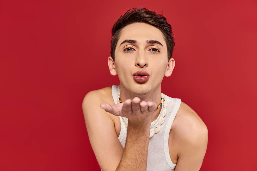 přitažlivý gay muž v bílém ležérní oblečení s doplňky foukání polibek na kameru na červeném pozadí - Fotografie, Obrázek
